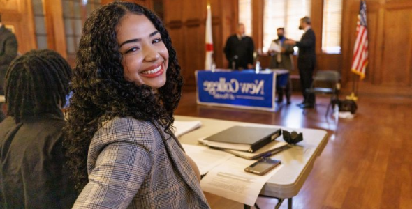 Smiling College age girl wearing a suit sits in beautiful wooden classroom where three teachers stand behind a table that has a large New College sign on it.